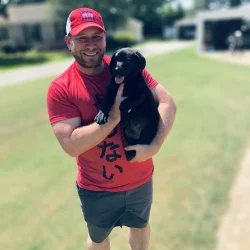 A man in a red shirt and cap holding a small black puppy outdoors.