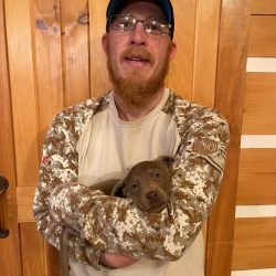 A man with a red beard and glasses holding a brown puppy against his chest.