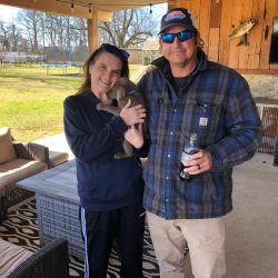 A woman holding a small puppy stands next to a man holding a bottle under a covered patio.