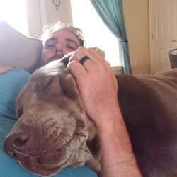 A man lying on a sofa rests his hand on the head of a sleeping brown dog.