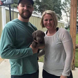 A man and a woman standing outside, holding a brown puppy.